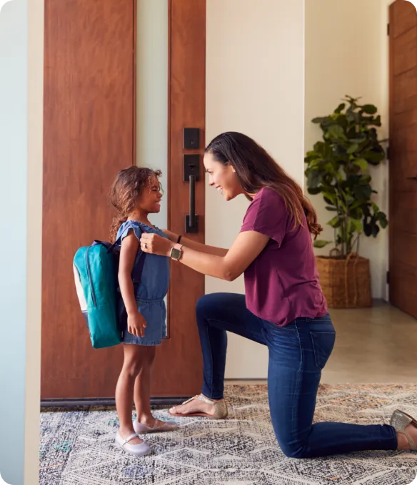 A mother kneels at the front door to help her young daughter with her backpack before the first day of school at Mosaic in Celina, Texas.