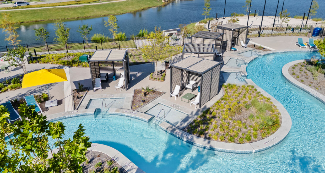 Aerial view of the resort-style lazy river at Mosaic, lined with shaded cabanas, lounge chairs, and lush landscaping. Beyond the pool area, a calm lake with a fishing dock sits in front of a row of new homes under a clear blue sky.
