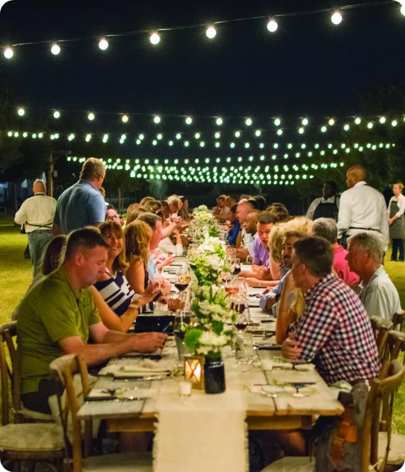 Neighbors share an outdoor farm-to-table dinner at a long, candlelit table under glowing string lights, a community event at Mosaic.