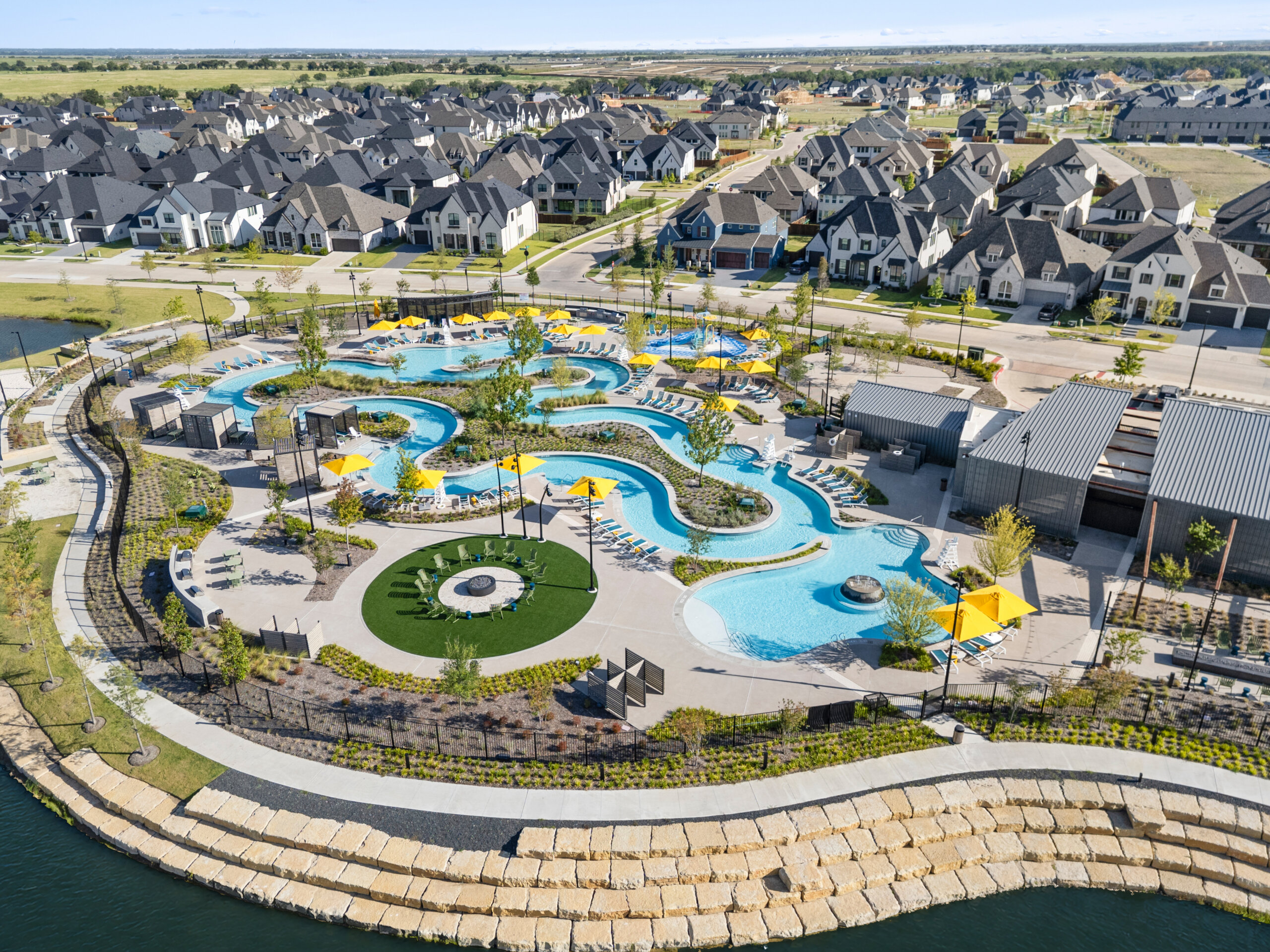 Aerial view of the River Amenity Complex at Mosaic in Celina, Texas. A winding lazy river is lined with yellow umbrellas, lounge chairs, and private cabanas, alongside a splash pad, a fire pit lawn, and the amenity center, with new homes in the background.